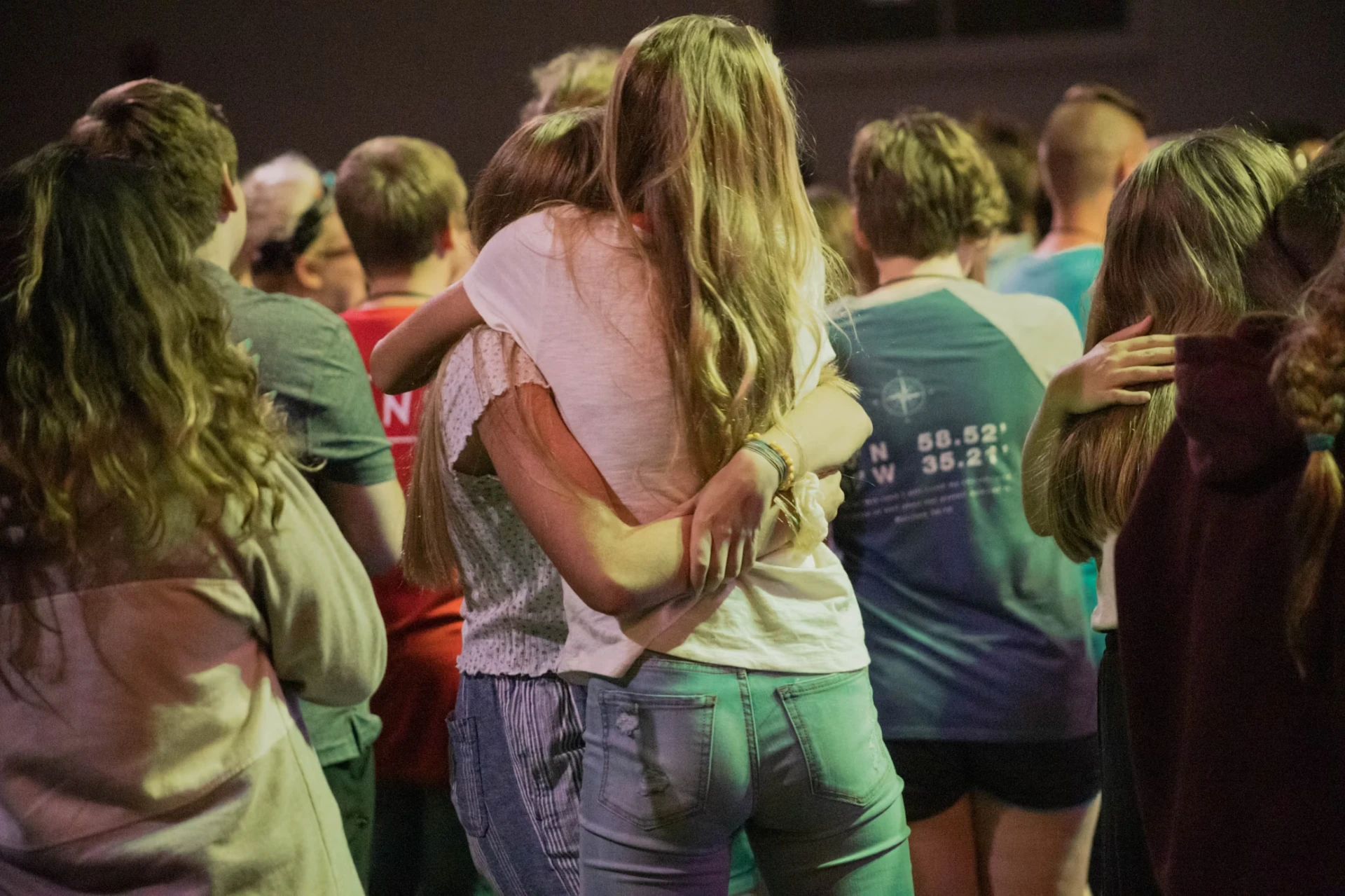 A group of young adults seen from behind, embracing each other while attending the "Youth Initiative" outdoor event at night.