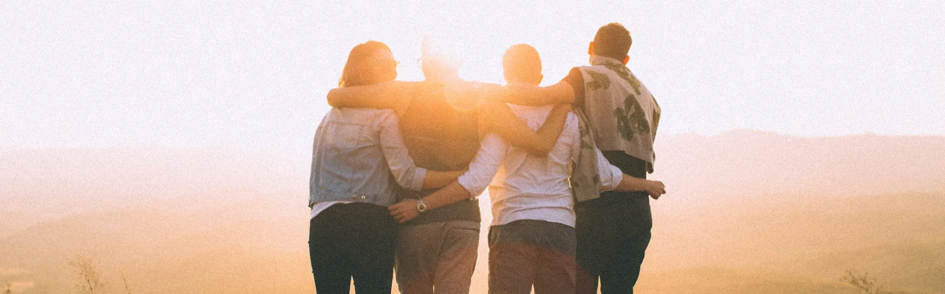 Four friends embracing during a visit while watching a sunset in a hazy landscape, symbolizing togetherness and friendship.