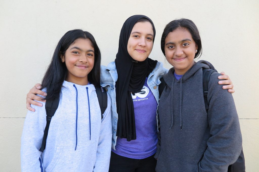 Three young girls smiling with arms around each other, standing in front of a plain wall for impact reports. One wears a hijab.