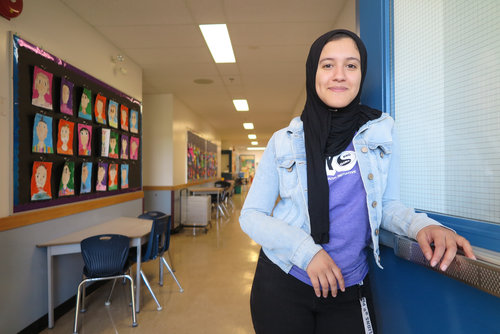 A young woman in a classroom, wearing a dacious denim jacket and scarf, smiles slightly while leaning on a window sill.