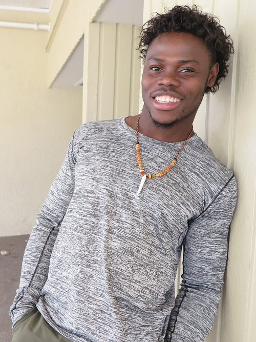 A young man with curly hair smiling at the camera, wearing a gray long-sleeve shirt and a beaded necklace, leaning against a pale yellow wall.