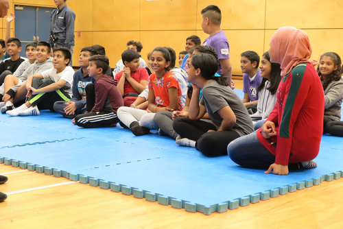 Children sitting on a blue mat in a gym, listening attentively, with adults standing in the background.