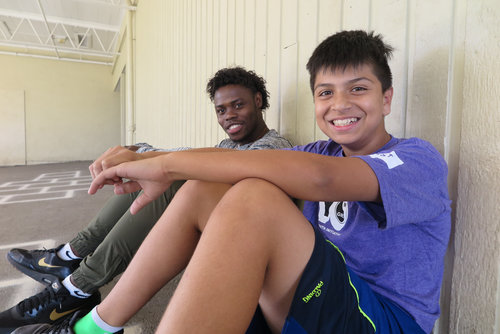 Two young men sitting and smiling against a wall, one with short curly hair and the other with a buzz cut, both dressed in sporty athletic attire.