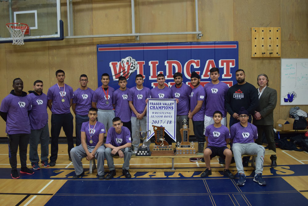 A group photo of a high school wrestling team with coaches, all wearing team shirts, standing behind a table displaying trophies and a championship banner. This content captures the celebratory moment effectively.