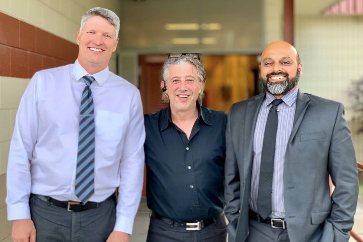 Three smiling men standing together outside a building in Kelowna, two in suits and ties and one in a casual black shirt.