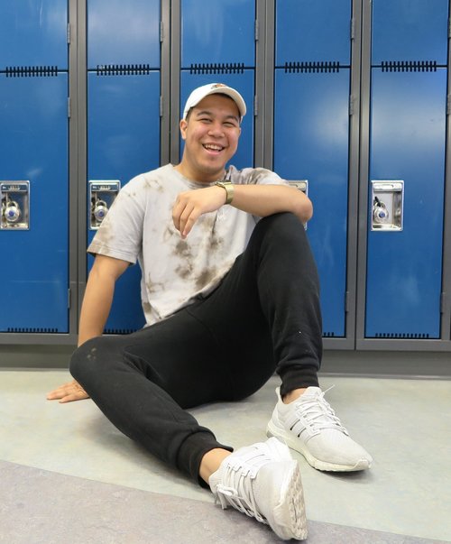 A young man smiling, sitting on the floor, leaning against blue lockers, wearing a stained white t-shirt and white sneakers.