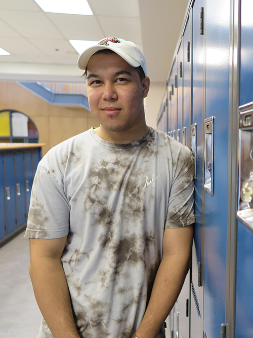 Young man in a tie-dye t-shirt and baseball cap standing by blue lockers, smiling gently at the camera, exuding a carefree vibe.