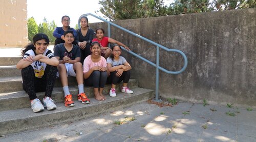 A group of eight diverse children, including Wahiba, smiling and sitting on steps next to a blue railing, outdoors on a sunny day.
