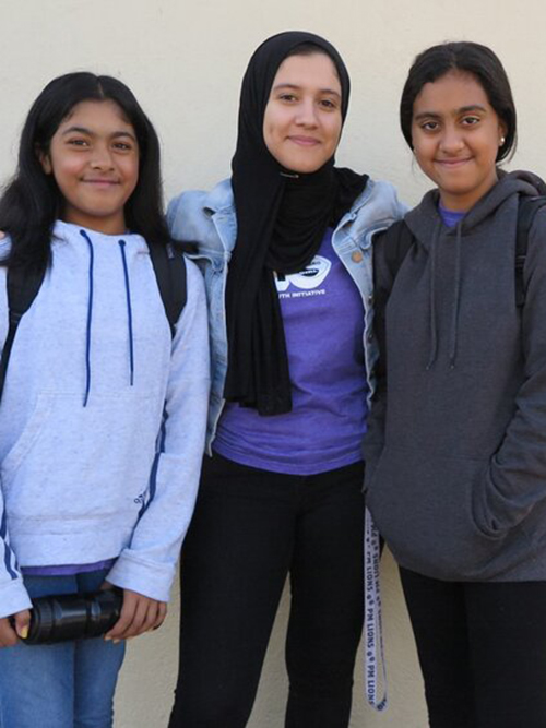 Three cheerful teenage girls, one wearing a hijab, are happily posing together against a light-colored wall.