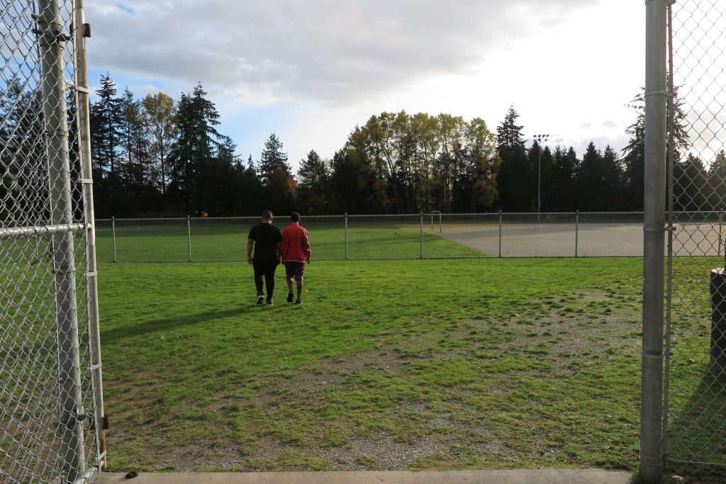 Two people walking towards a baseball field enclosed by a chain-link fence, with trees and clouds in the background as Billy waits at the entrance.