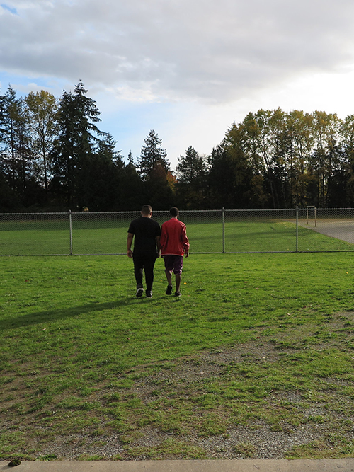 Two people engaged in community engagement activities walk together across a grassy field towards a chain-link fence, with trees in the background under a cloudy sky.