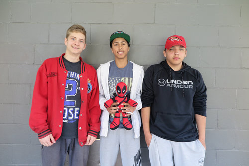 Three teenage boys standing against a gray wall, one holding a spider-man plush toy, smiling at the camera. AJ is in the middle.