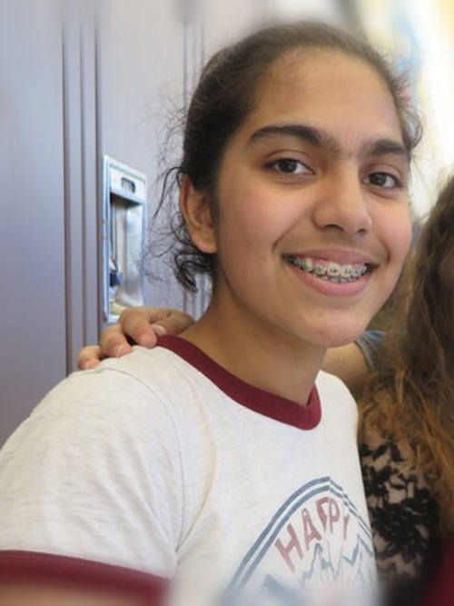 A smiling teenage girl with braces, wearing a white and burgundy t-shirt that reads