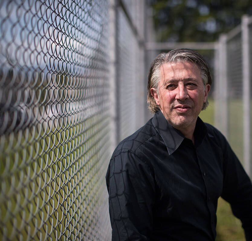 Man with gray hair, wearing a black shirt, leaning on a chain-link fence, smiling slightly at the camera for the 2019/2020 Impact Report.