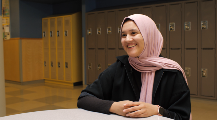 A young woman named Wahiba, wearing a pink hijab, sitting at a table, smiling and looking away, with blue lockers in the background.